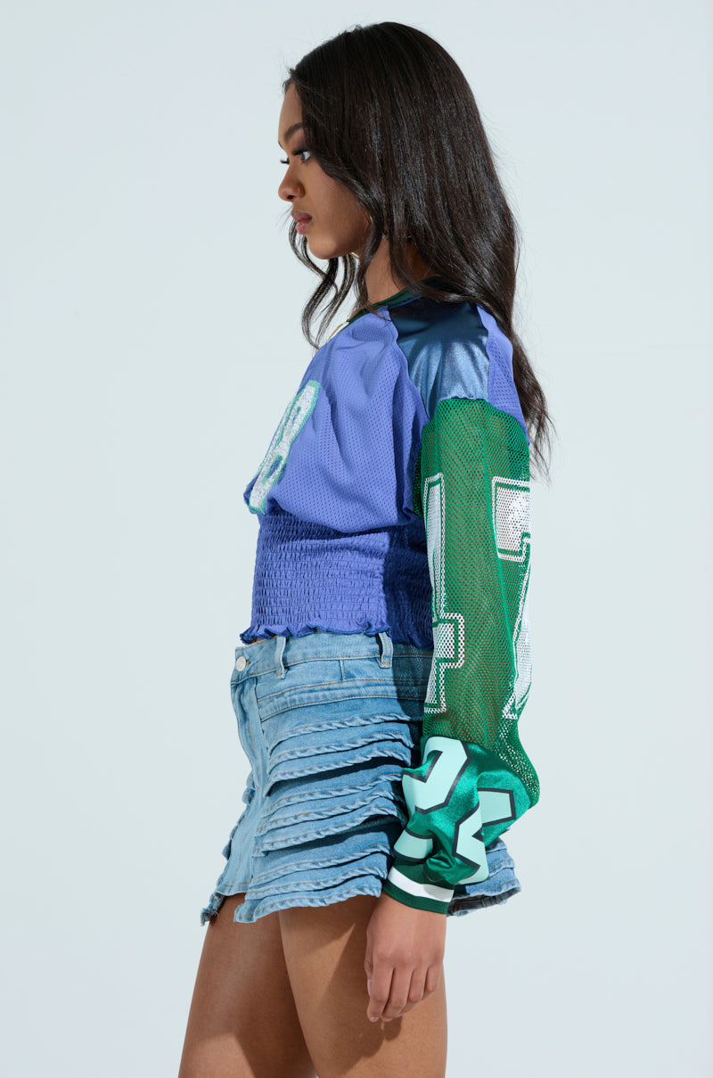 A person with long hair stands sideways, wearing the color-blocked MEXICO RUCHED JERSEY in blue and green shades, adorned with numbers. Light blue denim shorts with layered ruffles complement the look. The plain, light-colored background enhances the outfit's vibrancy.