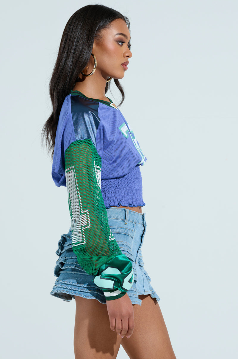 A woman in a color-blocked top with green sleeves and numbers, paired with denim shorts, stands in profile. Her long dark hair complements her large hoop earrings. The vibrant MEXICO RUCHED JERSEY adds flair to the light blue backdrop.
