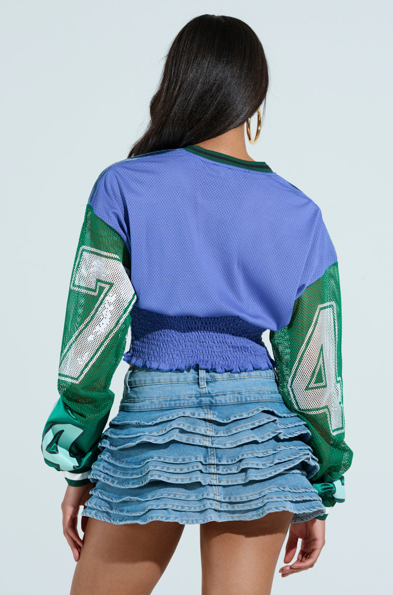 The person is wearing a MEXICO RUCHED JERSEY, a blue and green color-blocked top with the number 74 on the sleeves. They have long hair and are dressed in a layered ruffled denim skirt against a plain light blue background.