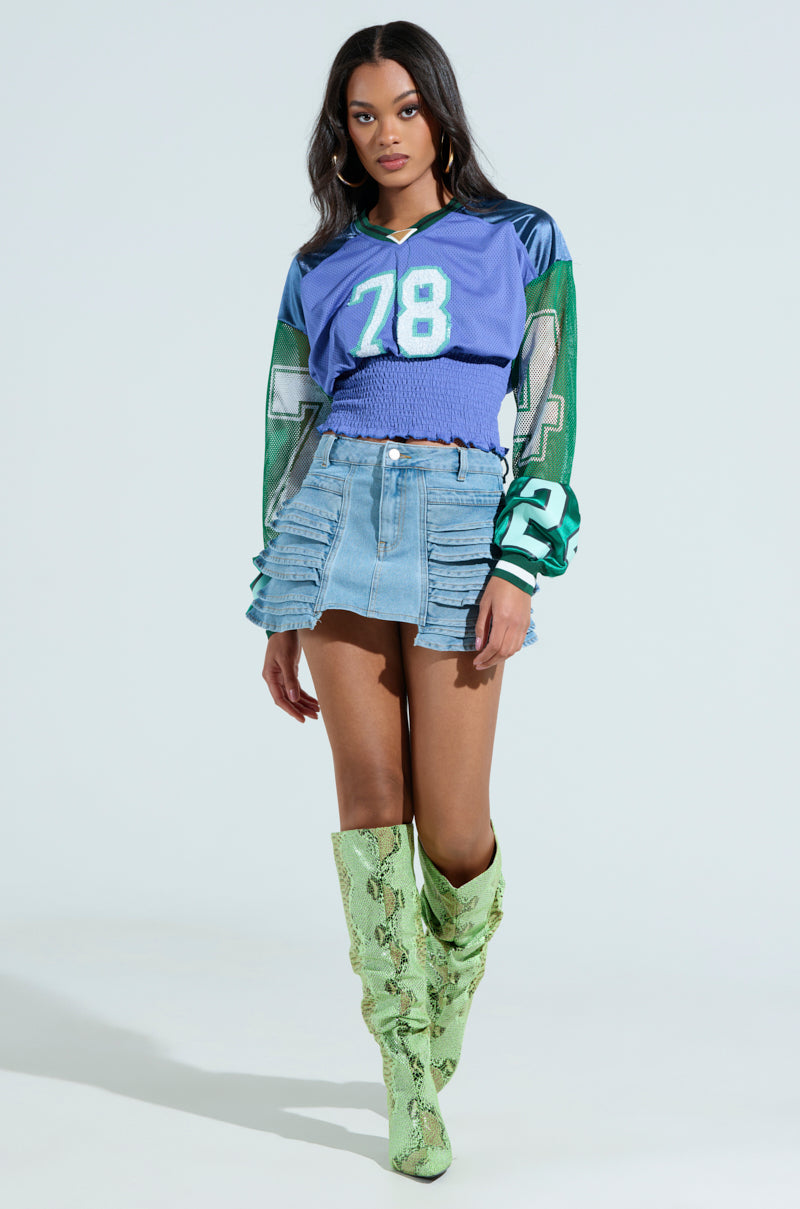 A woman poses in a MEXICO RUCHED JERSEY with the number 78, alongside a denim mini skirt and knee-high green snakeskin boots, against a plain light-colored backdrop.