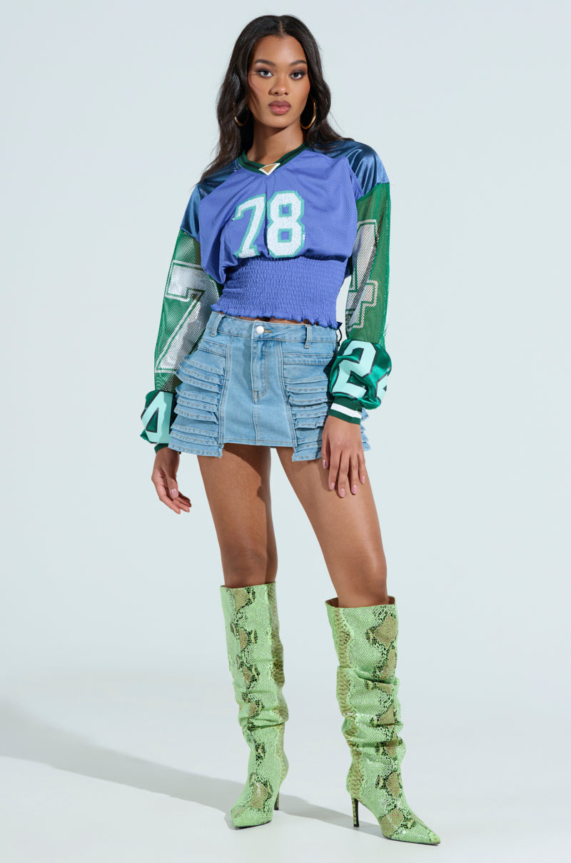 In a vibrant MEXICO RUCHED JERSEY top and denim mini skirt with ruffles, a woman confidently pairs her look with knee-high green snakeskin boots. The ensemble creates a striking contrast against the simple, light backdrop.