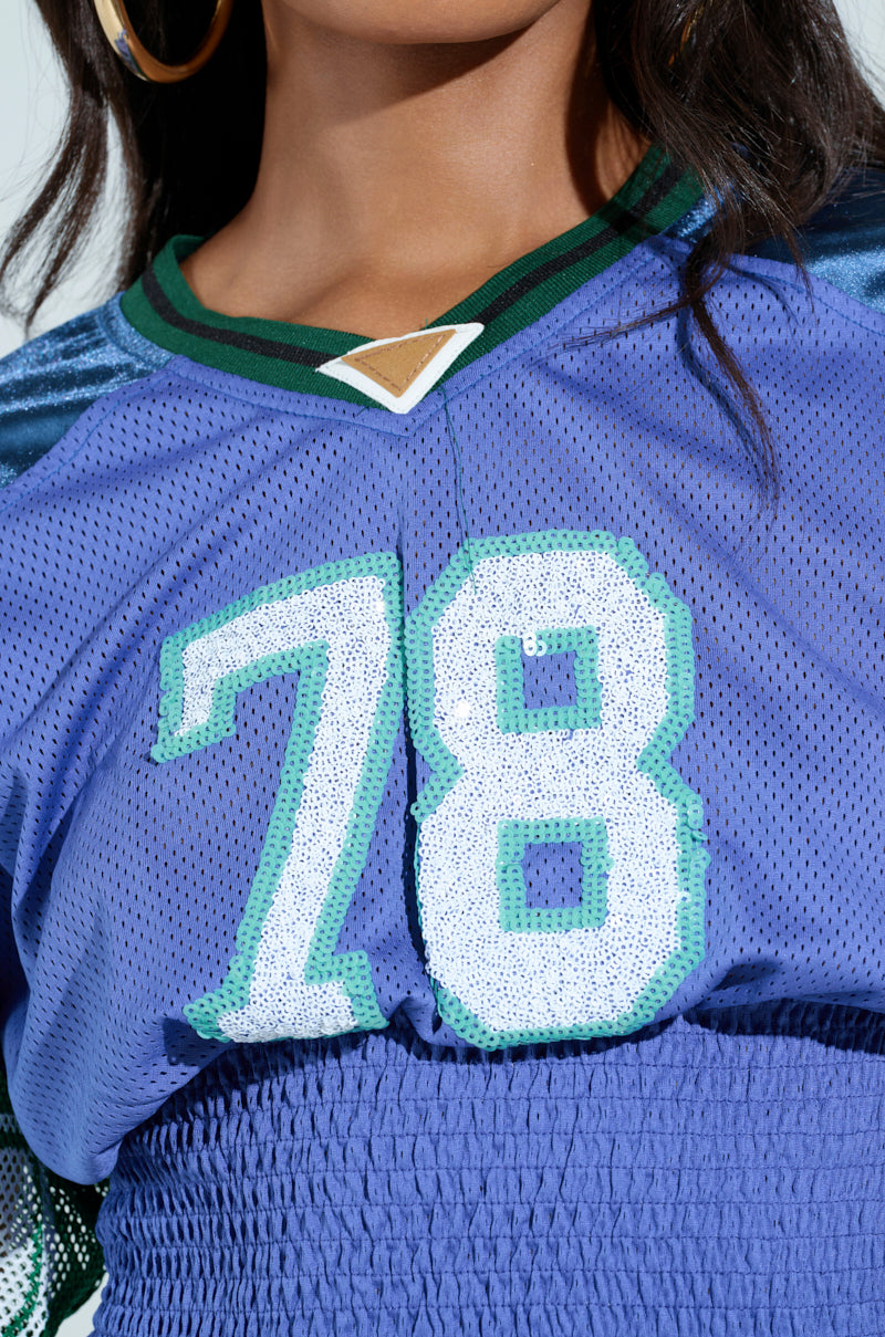 A person wears the MEXICO RUCHED JERSEY, a blue sports top with the number 78 in white and teal. It features color-blocked green and black trim around the neck and has a textured bottom half. A large hoop earring is visible on the left side.