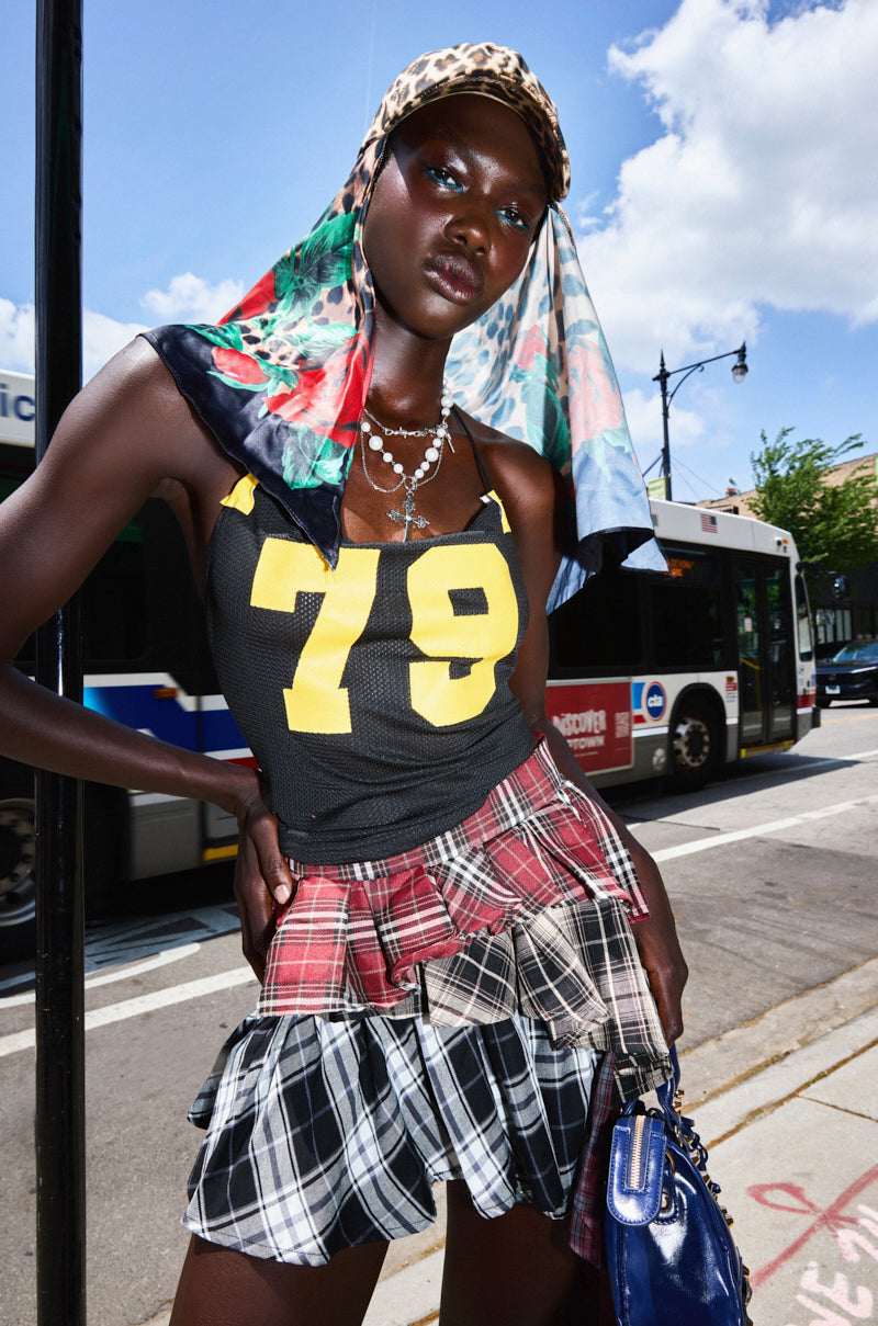 A stylish woman stands on a city street in the black GAME TIME DECISION JERSEY with yellow "78," paired with a red plaid skirt, layered necklaces, a patterned scarf, and cap. A bus and blue sky are visible behind her.