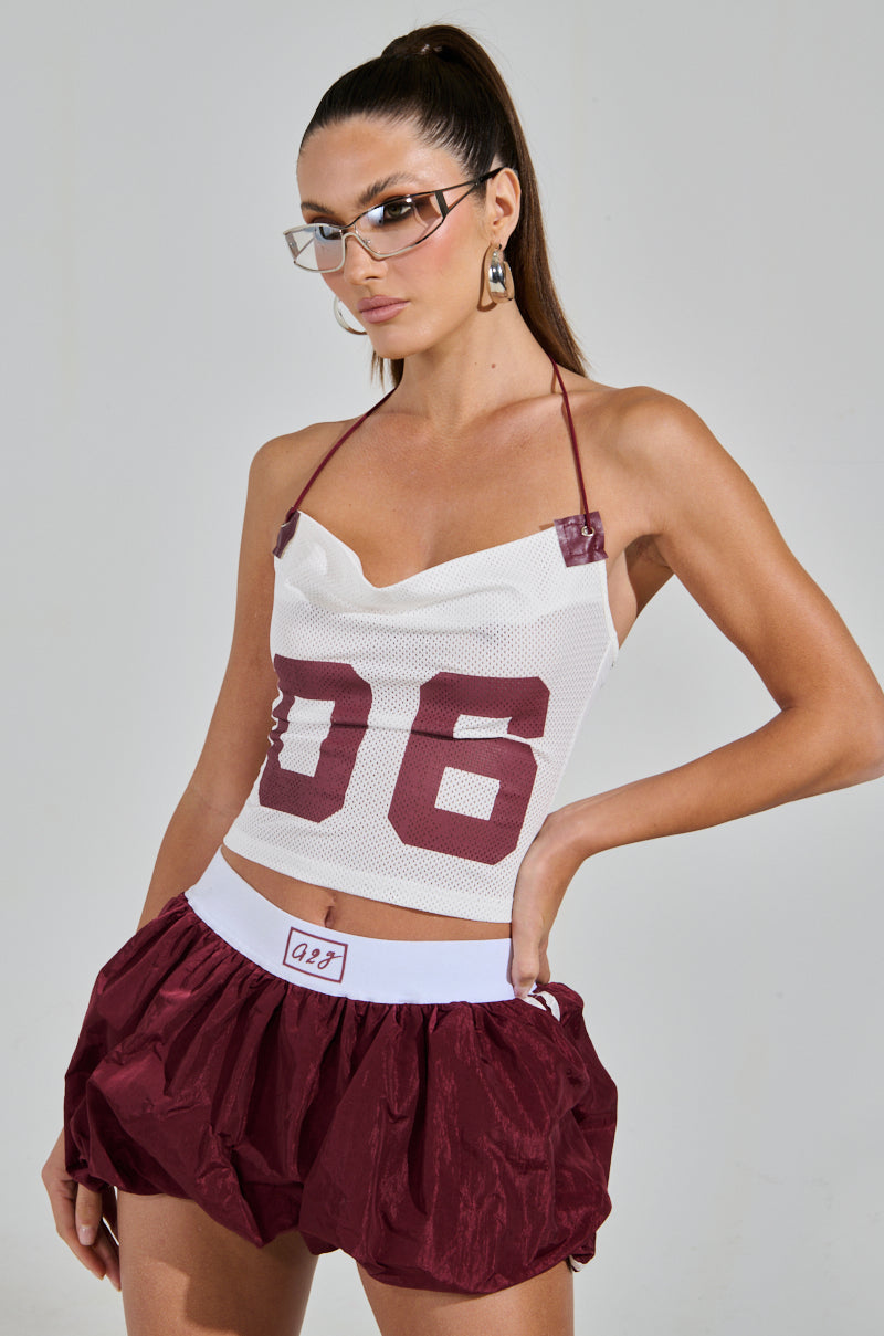 Sporting a chic look, a person with long hair in a ponytail and glasses confidently wears the stylish VARSITY SQUAD TANK with "06," paired with a voluminous skirt with a white waistband and large earrings for the perfect touch. 0