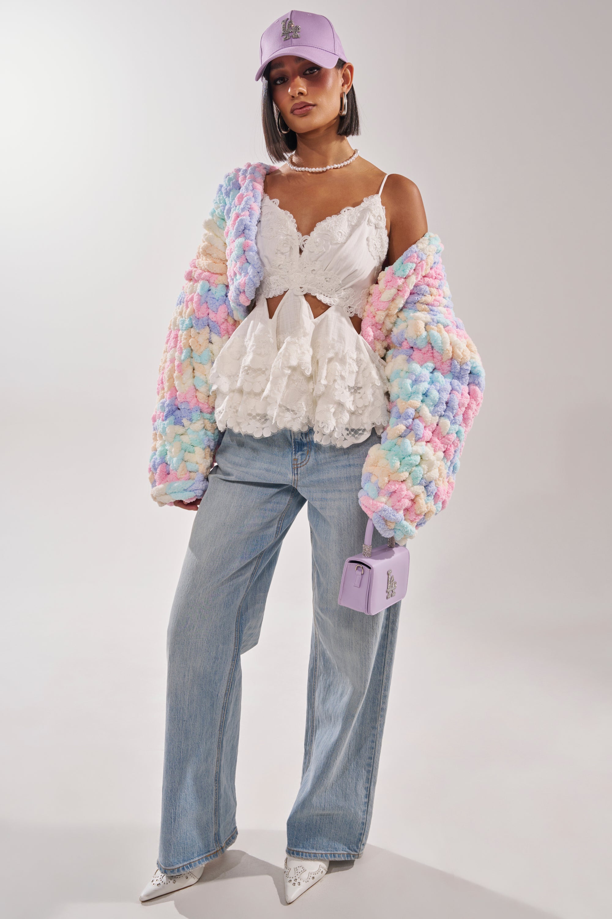 A woman wears the ALIA CHUNKY CHENILLE KNIT CARDIGAN in pastel, a white lace ruffle top, wide-leg jeans, white heels, a lilac baseball cap, and carries a small lilac handbag against a plain background. 9