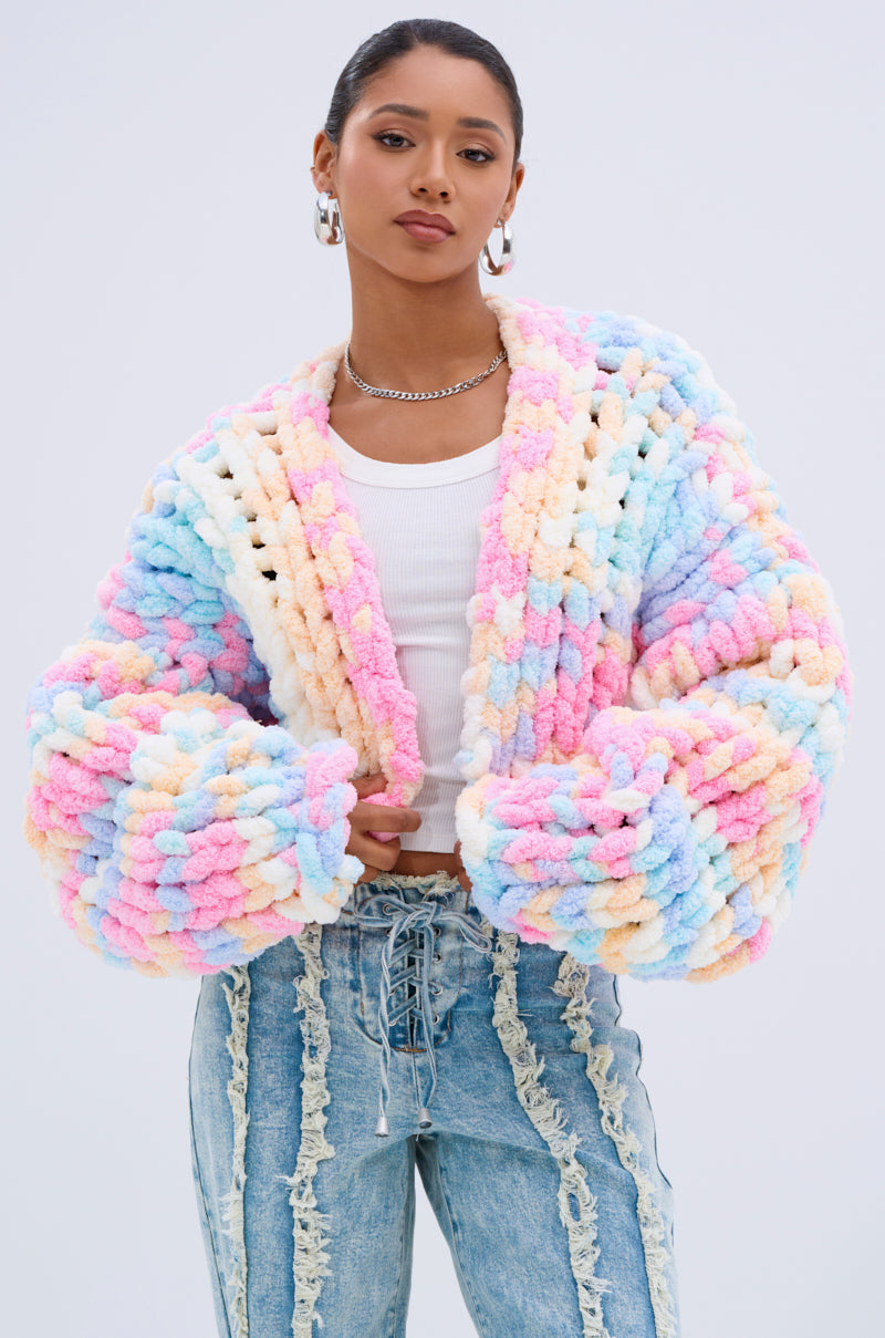 A woman confidently stands against a light background wearing the ALIA CHUNKY CHENILLE KNIT CARDIGAN in an oversized pastel style, layered over a white crop top and high-waisted distressed blue jeans.