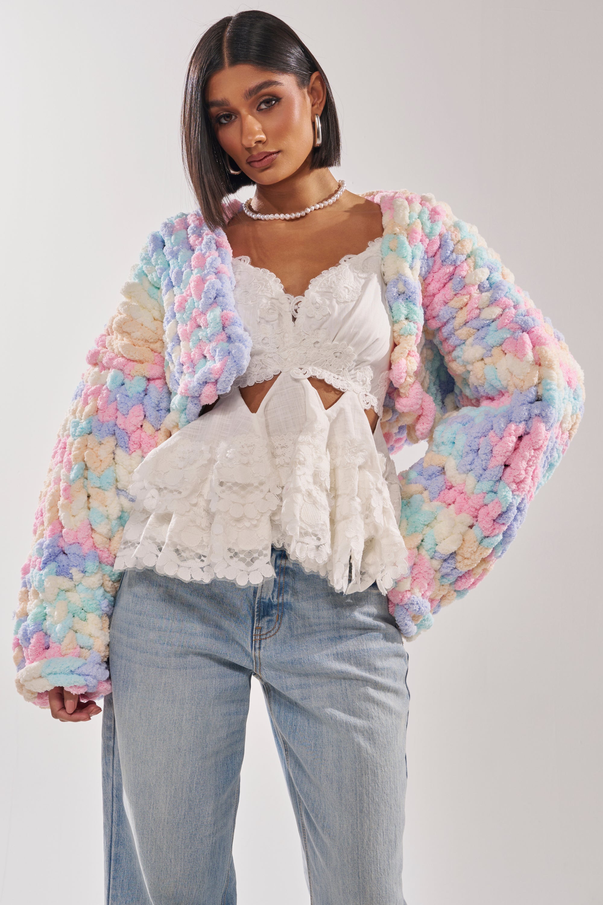Wearing the ALIA CHUNKY CHENILLE KNIT CARDIGAN in a pastel hue, a woman confidently poses against a plain background, paired with a white ruffled lace top and light-wash jeans. 1