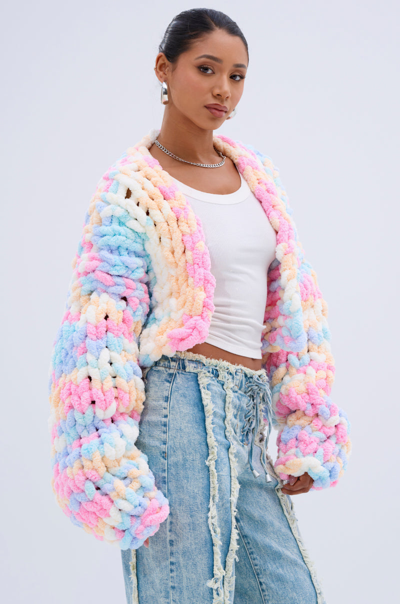 Wearing the ALIA CHUNKY CHENILLE KNIT CARDIGAN in a pastel oversized fit, paired with a white crop top and distressed high-waisted jeans, a person stands confidently against a plain light background.