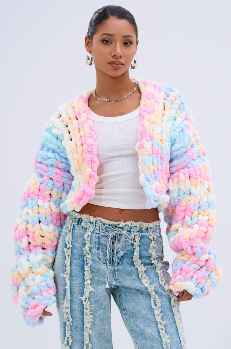 A woman stands confidently against a plain light background, wearing the ALIA CHUNKY CHENILLE KNIT CARDIGAN in an oversized pastel style over a white crop top and high-waisted, lace-up blue jeans.