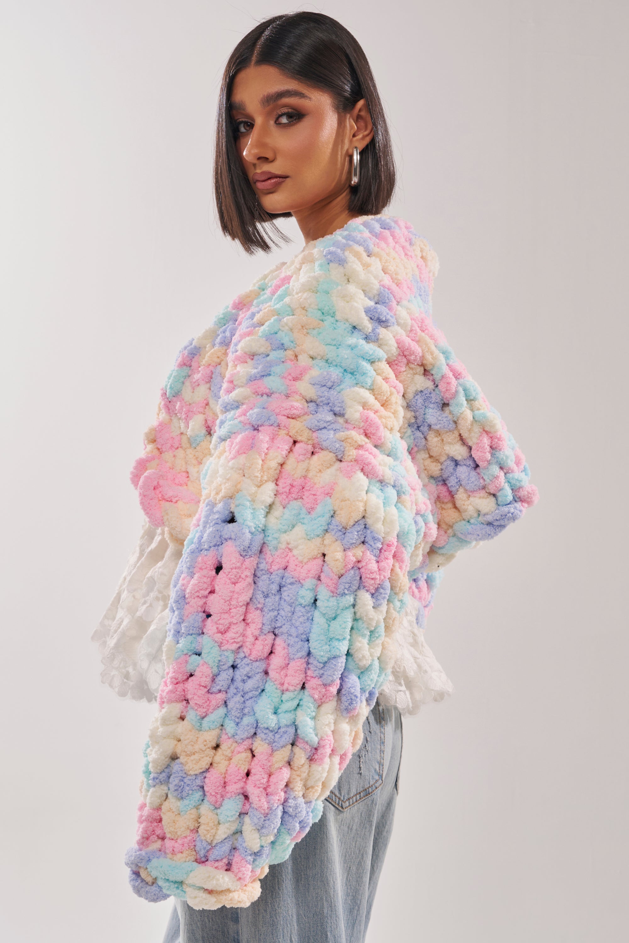 A woman with a sleek bob wears the ALIA CHUNKY CHENILLE KNIT CARDIGAN in pastel hues over a white top and light blue jeans, standing against a plain light background and glancing over her shoulder. 3