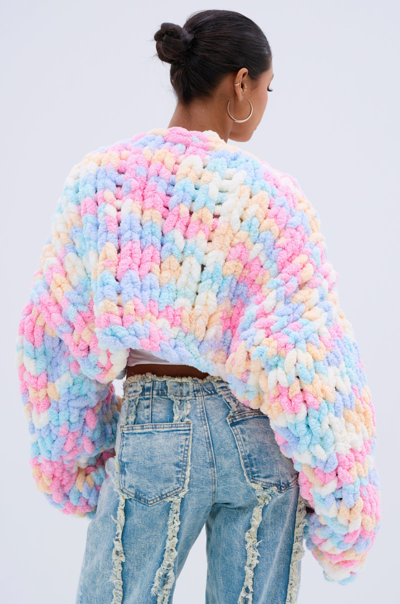 A woman with dark hair in a bun wears large hoop earrings, distressed high-waisted jeans, and the oversized fit ALIA CHUNKY CHENILLE KNIT CARDIGAN in a pastel colorway with chunky sleeves. She is facing away from the camera.