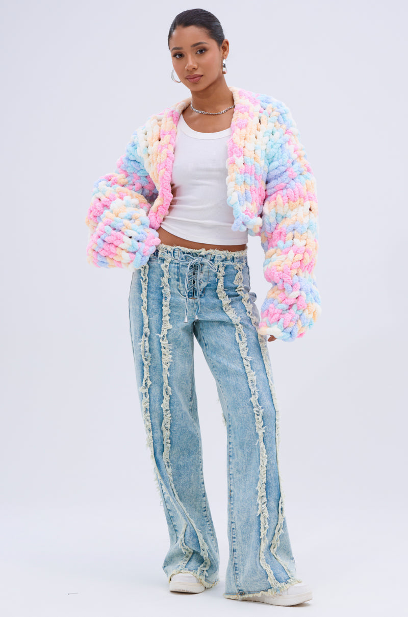 A woman stands against a plain background wearing the ALIA CHUNKY CHENILLE KNIT CARDIGAN in an oversized pastel style, paired with a white tank, light-wash wide-leg jeans with frayed seams, white sneakers, and hoop earrings.