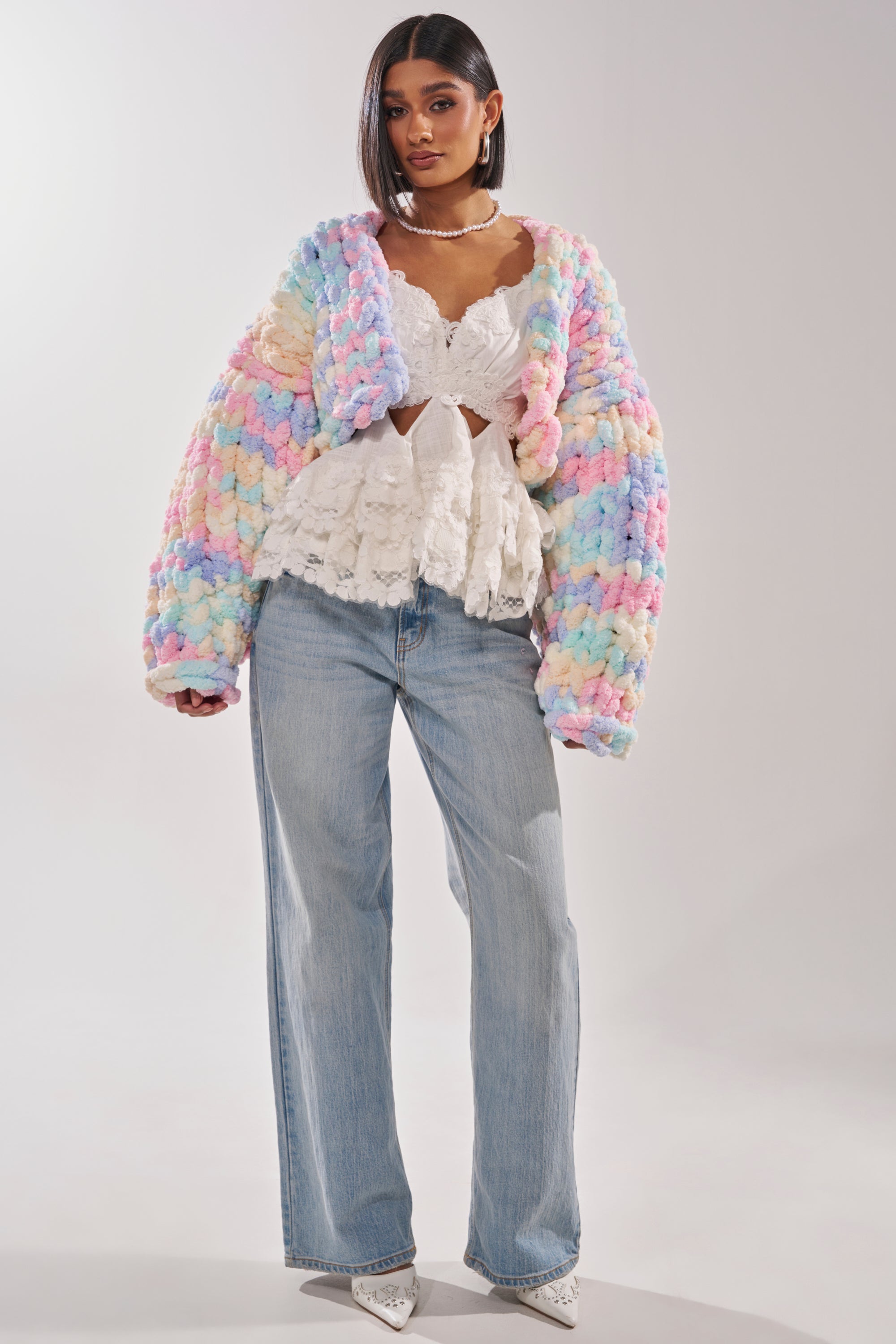 A woman stands facing forward against a plain light background, wearing an oversized ALIA CHUNKY CHENILLE KNIT CARDIGAN in pastel, a white lace peplum top, light blue wide-leg jeans, and white pointed shoes. 5