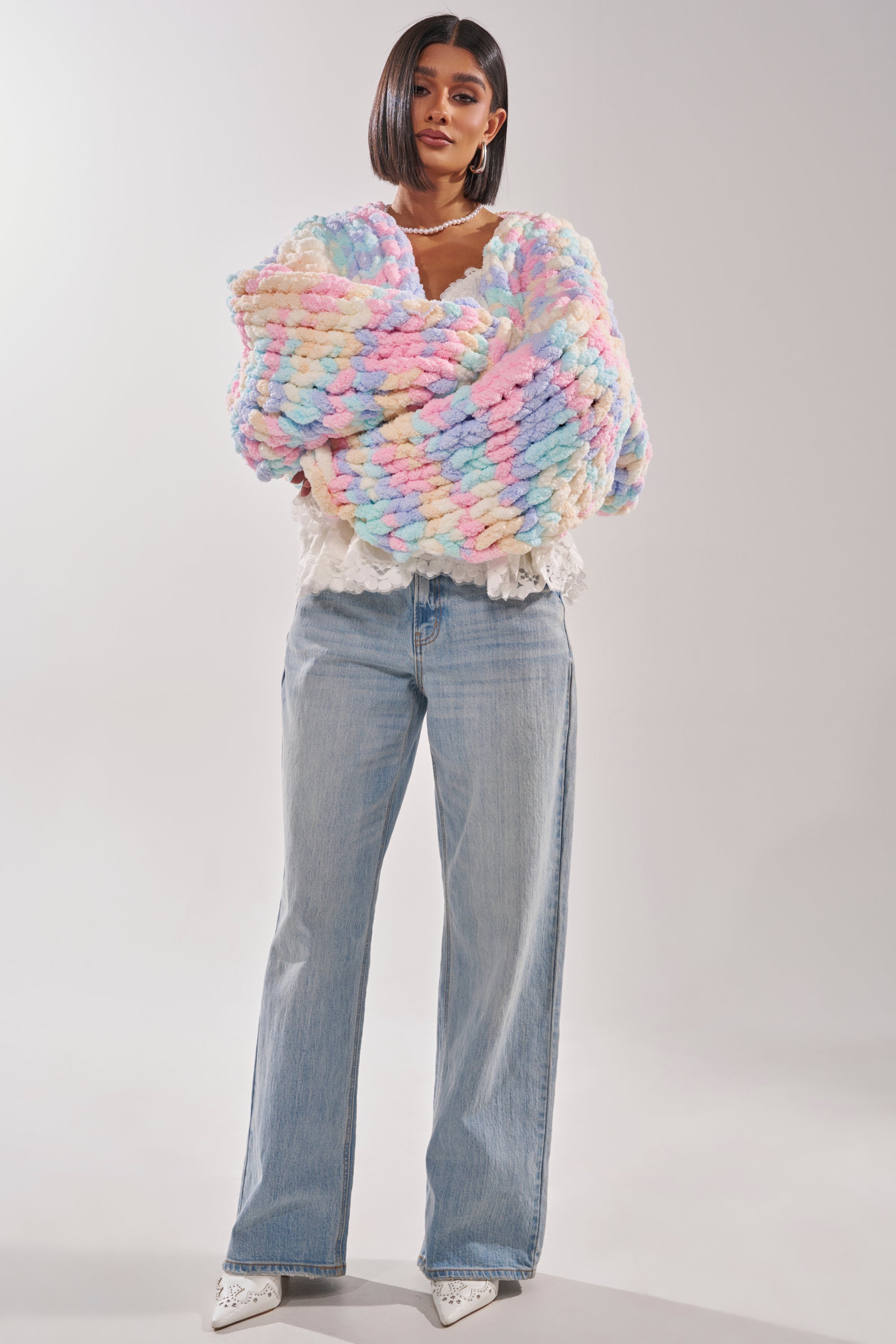 A woman with straight, shoulder-length dark hair stands facing forward, wearing the ALIA CHUNKY CHENILLE KNIT CARDIGAN in a pastel hue, paired with a white lace blouse, light-wash wide-leg jeans, and white shoes. 6