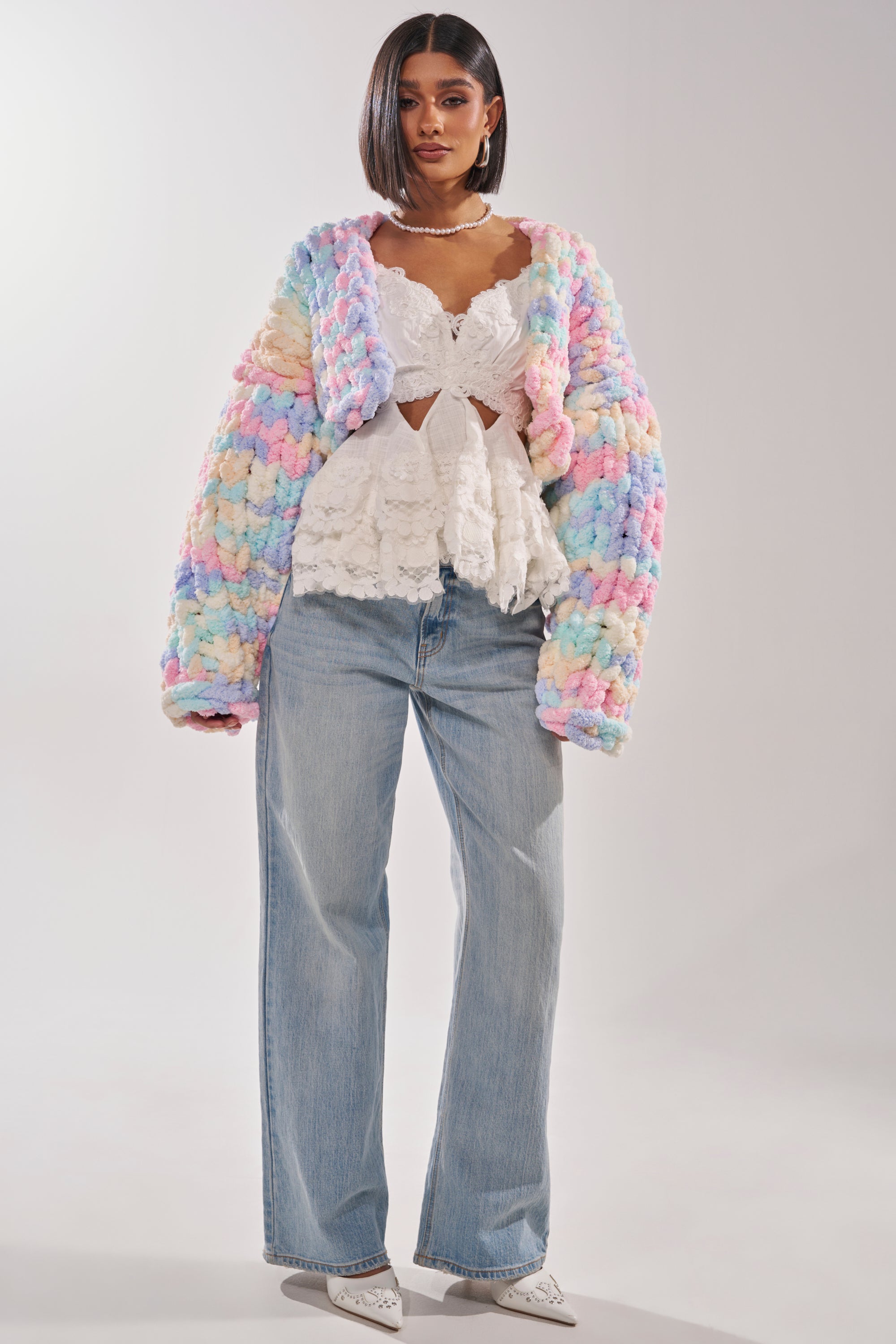 A woman poses in light blue wide-leg jeans, a white lace top, and the ALIA CHUNKY CHENILLE KNIT CARDIGAN. She finishes the outfit with white pointed shoes and a pearl necklace, standing against a neutral background. 7