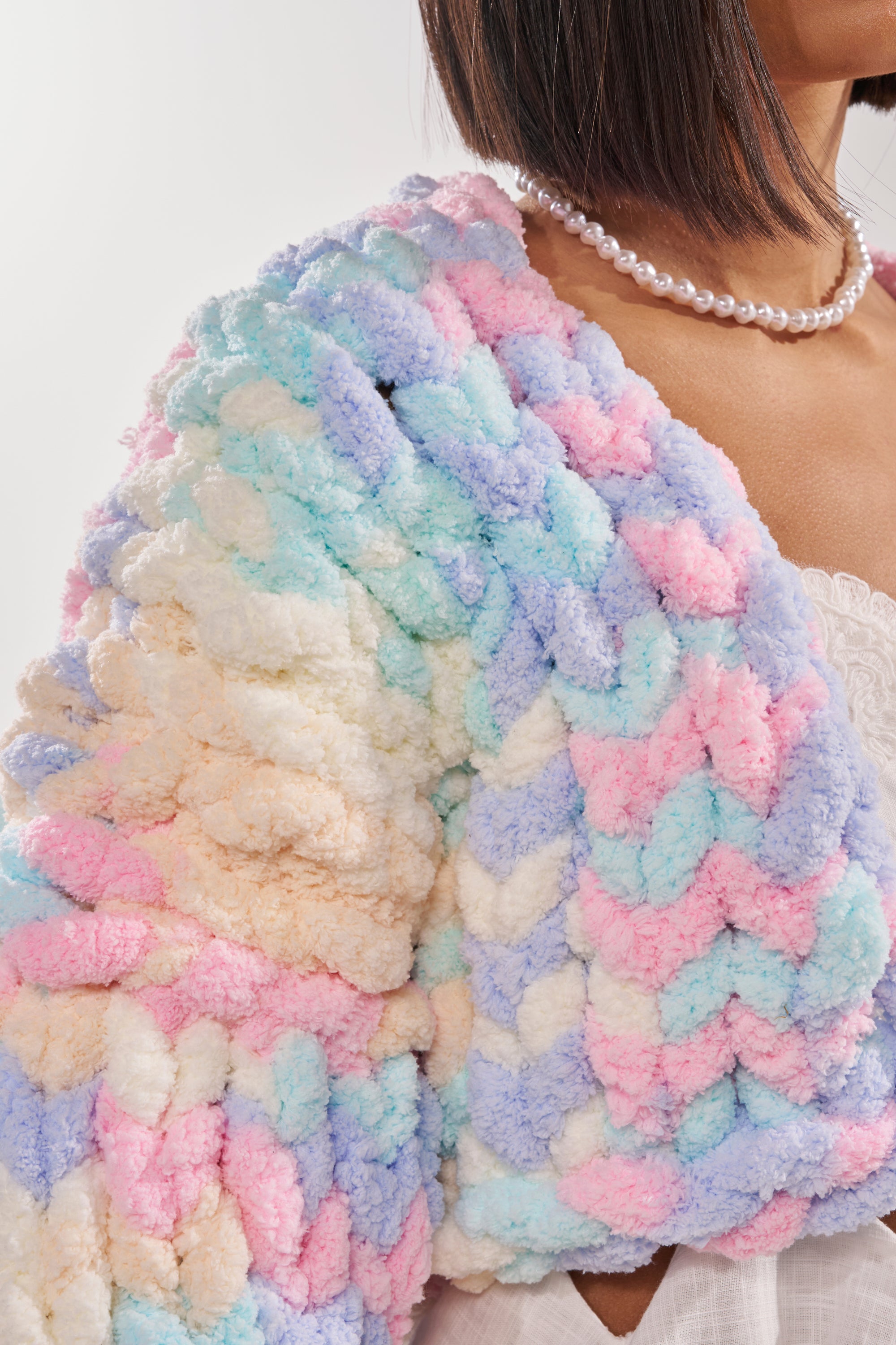 Close-up of a person wearing the ALIA CHUNKY CHENILLE KNIT CARDIGAN in oversized pastel pink, blue, and cream, styled with a pearl necklace and a white lace top. Only the lower face and torso are visible. 8