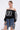 A person poses confidently in the UNKNOWN JERSEY, a sporty-chic black off-shoulder top with the number 83, paired with a statement necklace, hoop earrings, and a denim strip mini skirt against a plain white background.