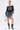 A woman poses in the UNKNOWN JERSEY—a sporty-chic, off-shoulder black top with 83—plus a patchwork denim skirt with lace trim, plaid heeled boots with red and blue scrunchy accents, and a small metallic square handbag.