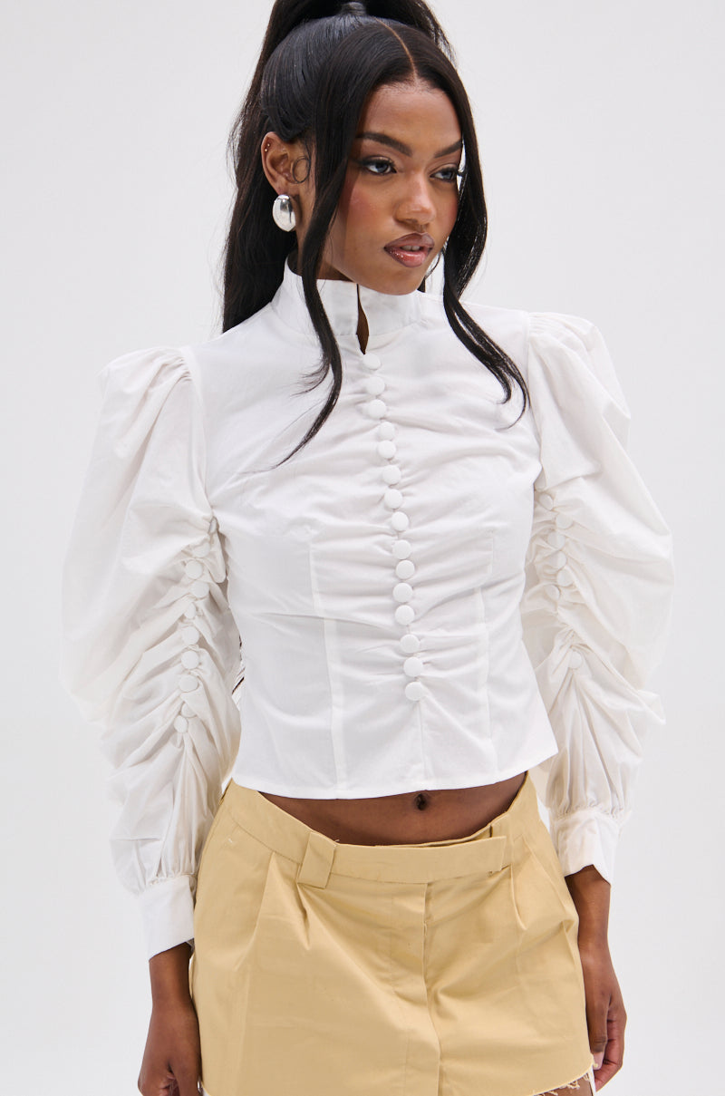 Wearing the GIVE ME LOVE WHITE BLOUSE with peasant sleeves and metallic back zipper, a person pairs it with a beige skirt, styled with long black hair in a high ponytail, hoop earrings, all set against a plain white background.
