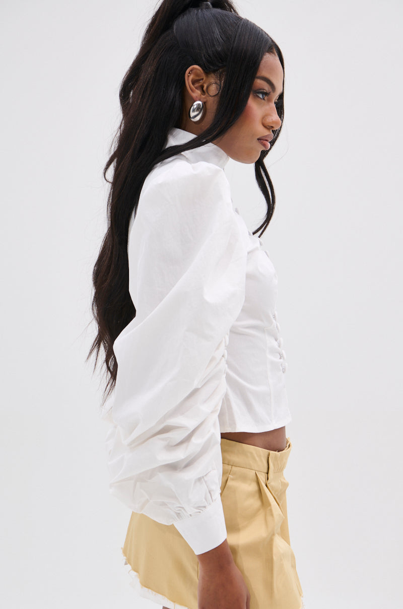 A woman with long, dark hair models a GIVE ME LOVE WHITE BLOUSE with peasant-style sleeves and beige shorts. She wears large silver earrings and poses against a plain white background, showcasing the blouse's metallic back zipper.