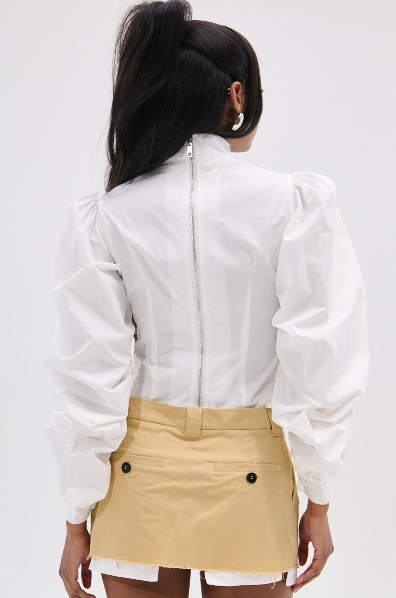 Rear view of a person wearing the GIVE ME LOVE WHITE BLOUSE with long dark hair, featuring peasant-style sleeves and a high neck, paired with a short beige skirt with black buttons. The back zipper is metallic, set against a plain white background.