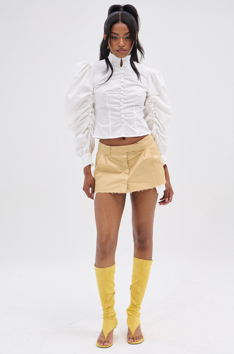 Against a plain background, someone wears the "GIVE ME LOVE WHITE BLOUSE" featuring peasant-style sleeves, paired with a short beige skirt with a metallic back zipper. Knee-high yellow boots complete the look, and their hair is in a high half-updo.