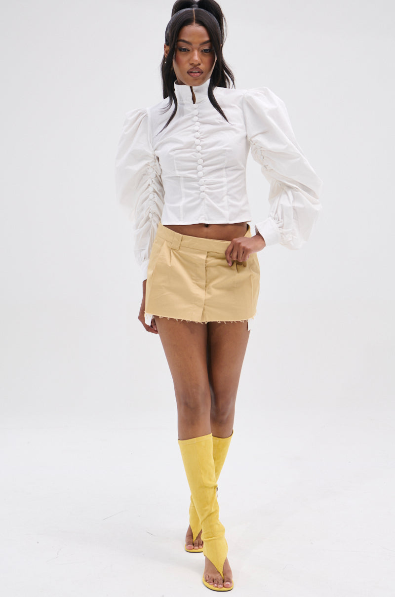A person in the "Give Me Love" white blouse with peasant-style sleeves and a high collar is paired with a beige mini skirt and yellow knee-high open-toe boots featuring metallic back zippers, standing against a light background.