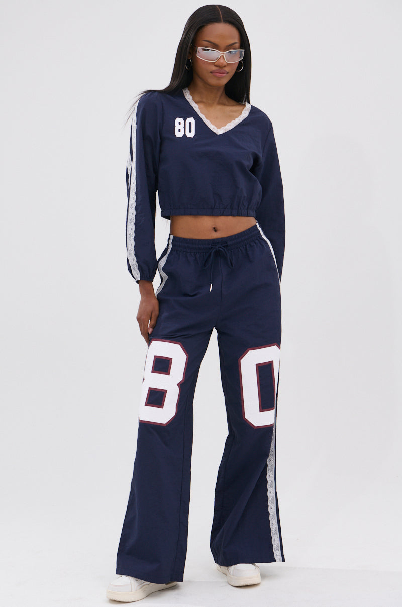 A woman wearing the LACE TRIM NYLON TRACK PANT as part of a navy blue athleisure two-piece with white and gray stripes, number "80" on the top, white sneakers, and clear-framed glasses stands against a plain white background.
