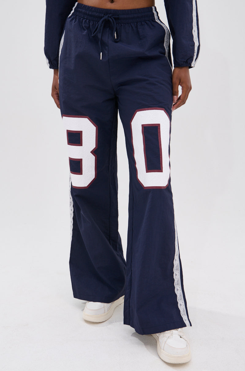 Wearing the LACE TRIM NYLON TRACK PANT—high-rise, wide-leg navy pants with bold white and red "80" on each leg—paired with white sneakers for an athleisure look. Only the lower half of the body is visible.