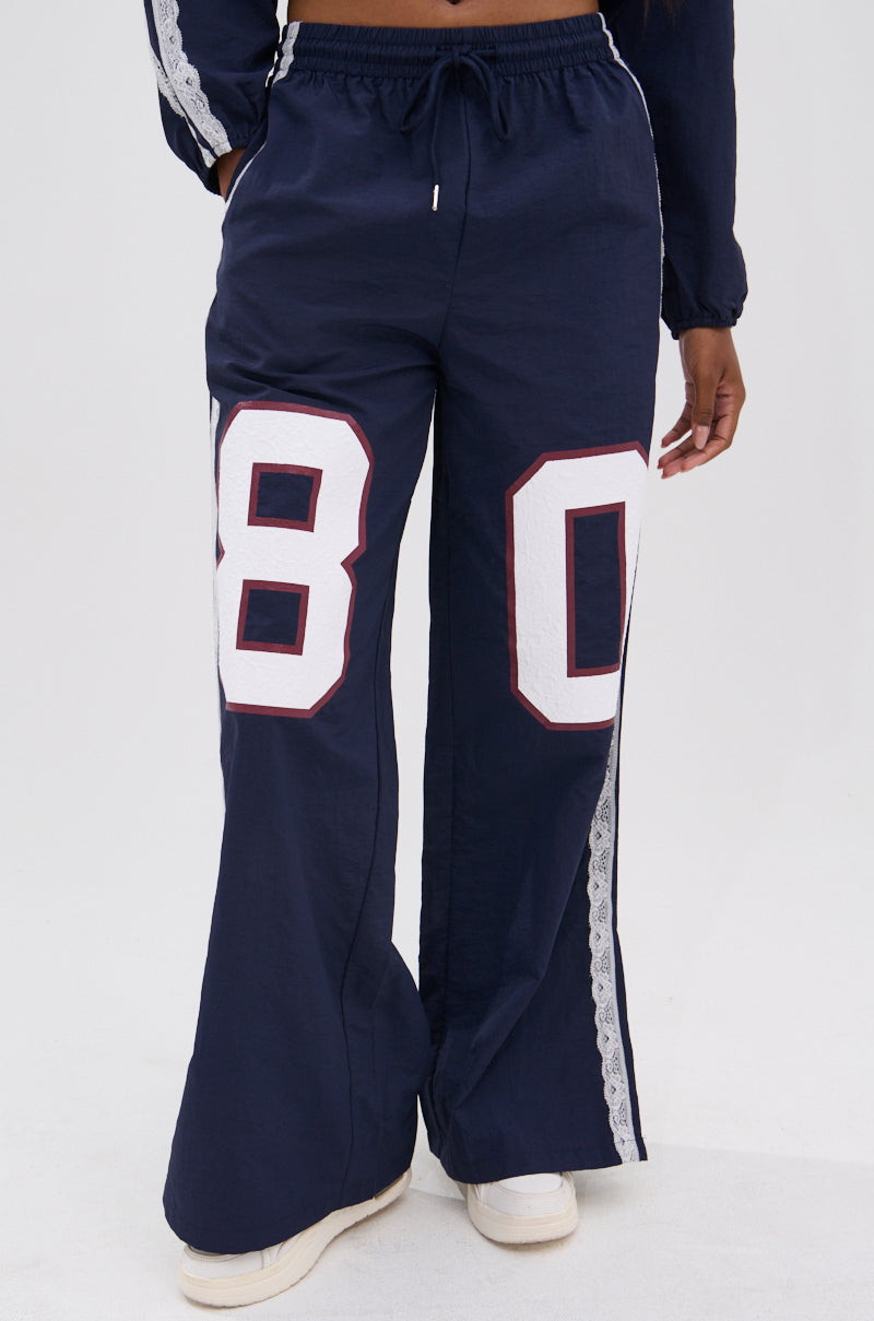 Someone is seen wearing LACE TRIM NYLON TRACK PANT featuring large white and red "80" numbers, white side stripes with lace trim, paired with white sneakers, standing against a plain background.
