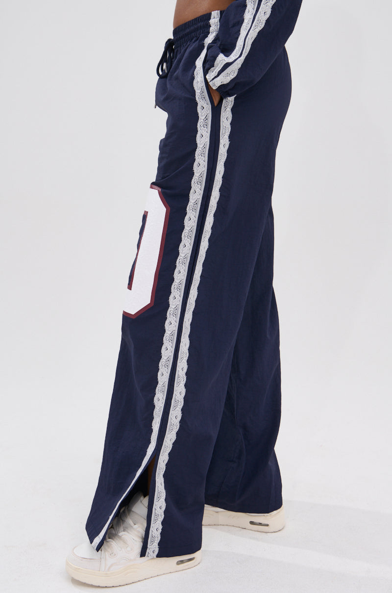 A person wears high-rise wide-leg LACE TRIM NYLON TRACK PANT in navy with white lace trim along the sides, a large bordered white patch, side slits revealing white sneakers for an athleisure look.