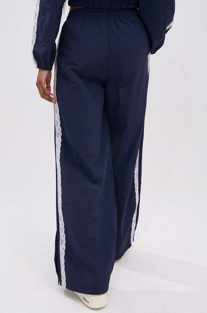 A person, shown from the back, models the LACE TRIM NYLON TRACK PANT—high-rise wide-leg pants with a white lace side stripe—paired with white shoes, all set against a crisp white background.