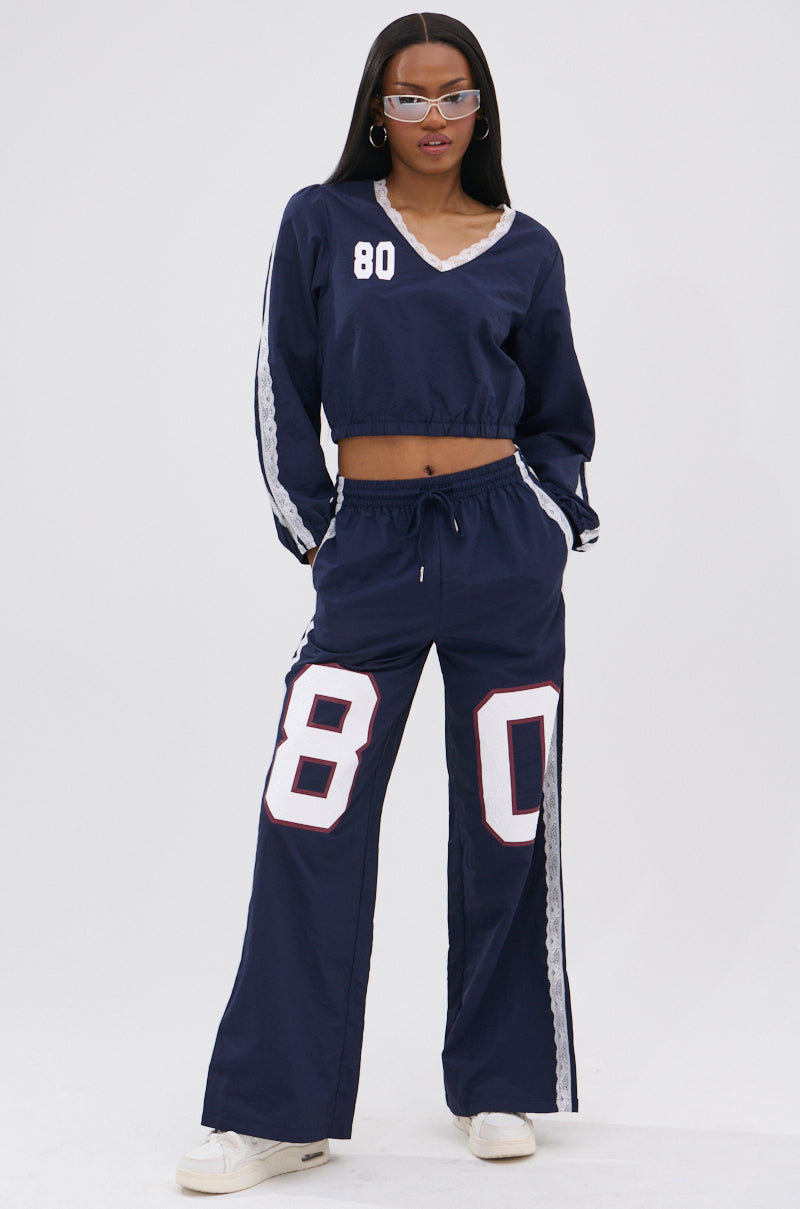 A woman stands against a plain white background wearing LACE TRIM NYLON TRACK PANT paired with a navy blue top featuring white "80" numbers, silver-rimmed sunglasses, hoop earrings, and white sneakers.