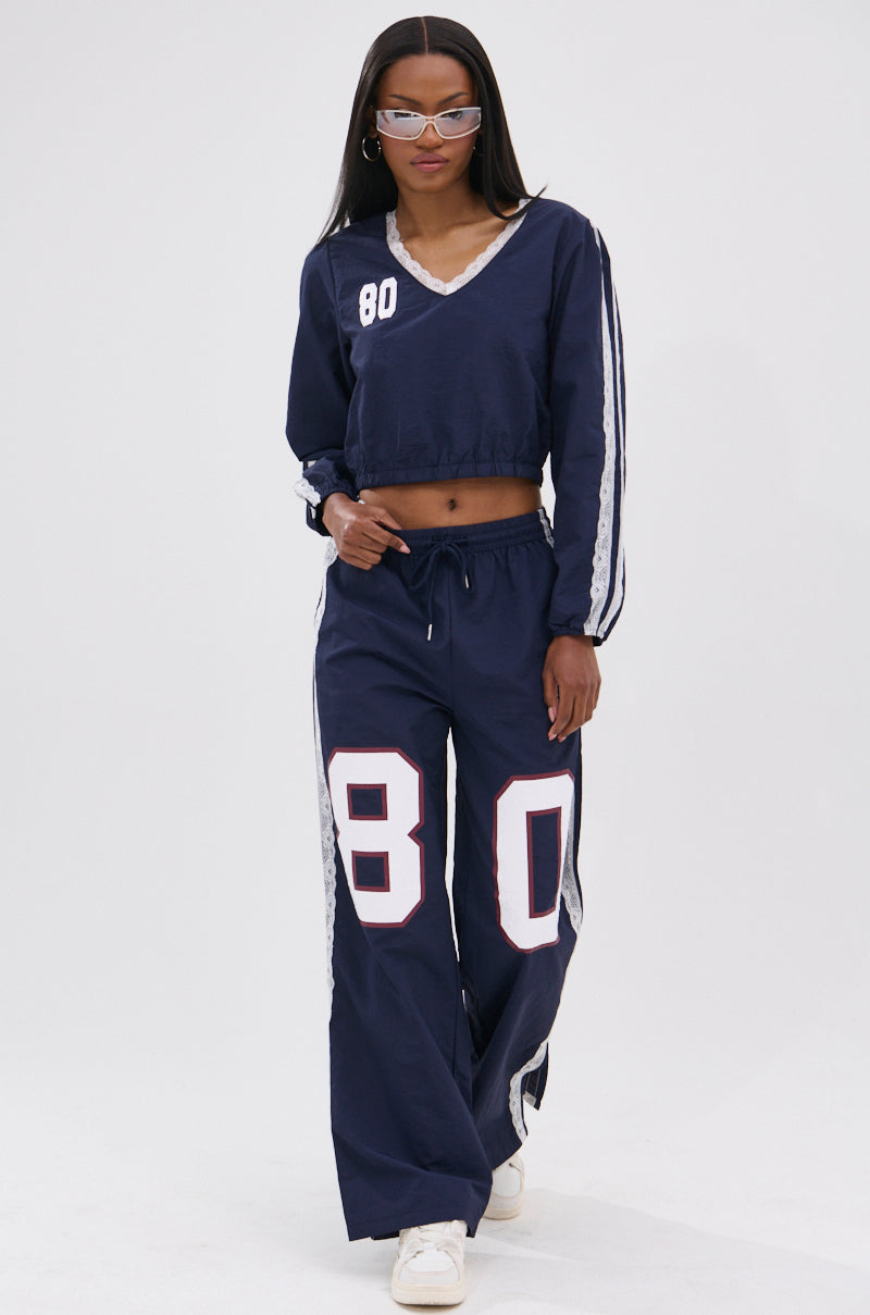 A woman walks confidently against a plain background in the LACE TRIM NYLON TRACK PANT, part of a navy athleisure two-piece featuring white and light blue accents, the number 80 on both top and high-rise wide-leg pants, with white sneakers and clear-frame glasses.