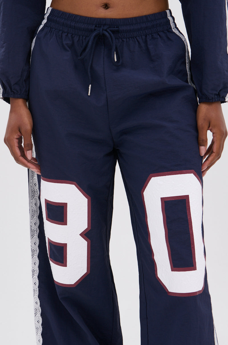 Someone wears the LACE TRIM NYLON TRACK PANT in navy with white lace stripes and "80" prints on the front. Only the lower torso and upper legs are visible, creating a sporty athleisure look.