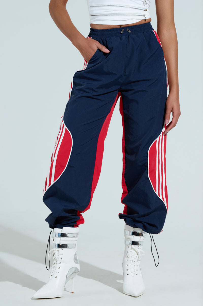 Dressed in the GAME TIME TRACK PANT with navy blue and red/white stripe details, a person stands hand-in-pocket, highlighting the color-blocked design. They pair the pants with white heeled boots and a sleeveless top against a light gray backdrop.