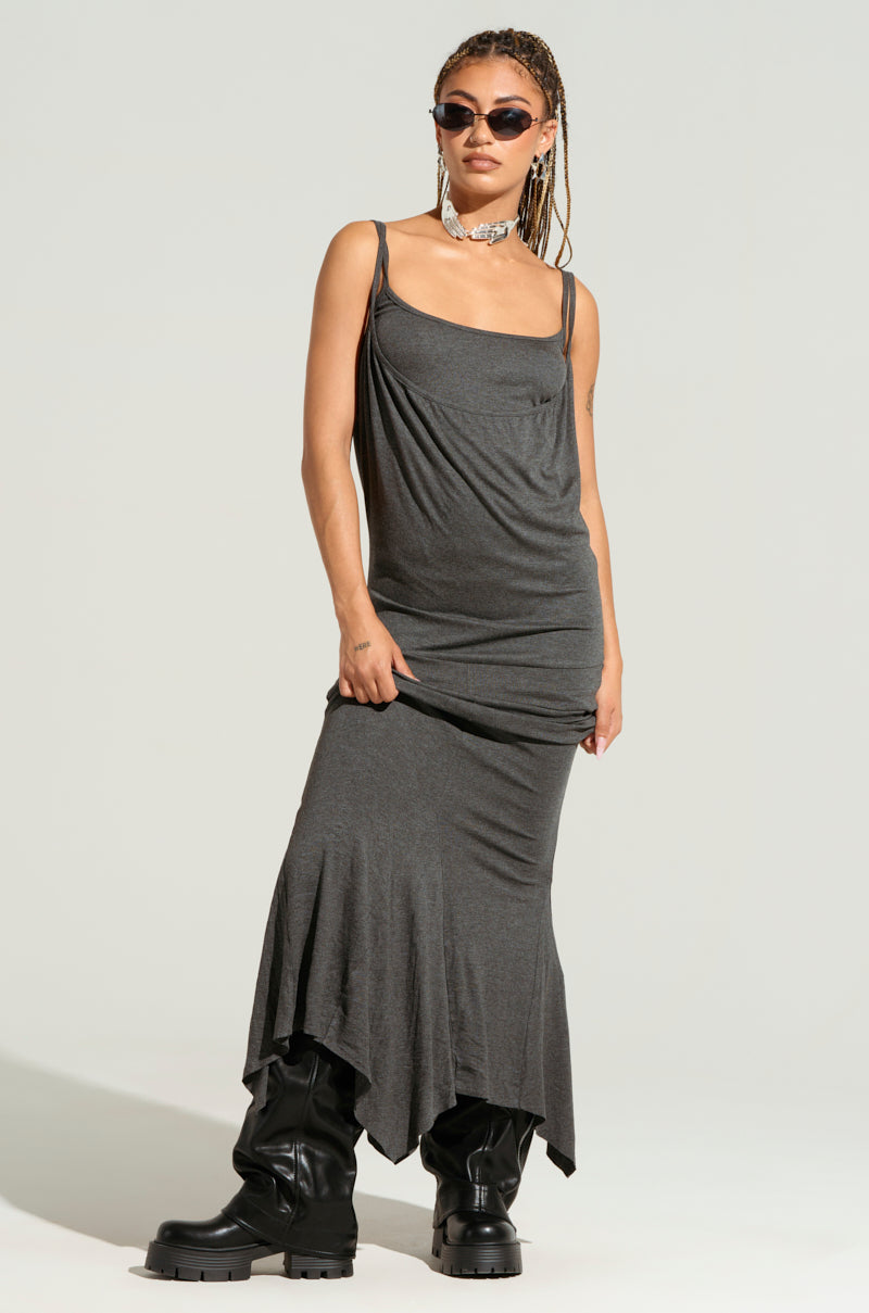 A person confidently poses in the WYNNIE KNIT MAXI DRESS SET, a super stretchy gray asymmetrical dress paired with black boots. They have braided hair, sunglasses, and a choker necklace. The plain backdrop highlights the stylish bodycon silhouette of their outfit.