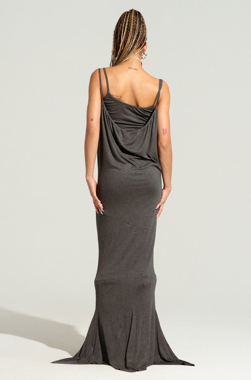 A person with braided hair stands facing away, wearing the WYNNIE KNIT MAXI DRESS SET in a deep gray shade, featuring a draped back design. The dress is crafted from an ultra-stretchy fabric that ensures comfort while contouring to a bodycon silhouette. The flowing hem sweeps the ground, revealing tattoos on their right shoulder and middle left arm.