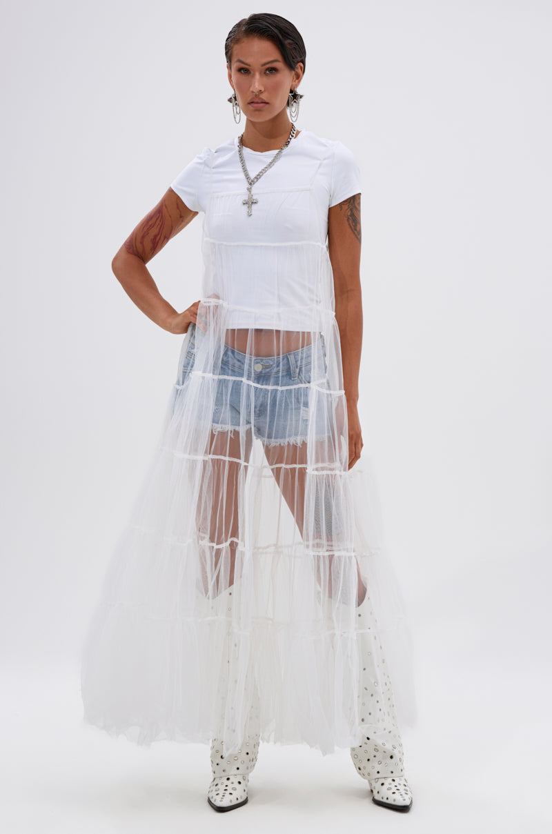 A person stands confidently in the PERFECT FOR LAYERING TULLE MAXI DRESS IN WHITE over a white tee and denim shorts, paired with studded knee-high boots, silver jewelry, and a cross necklace against a plain white background. 0