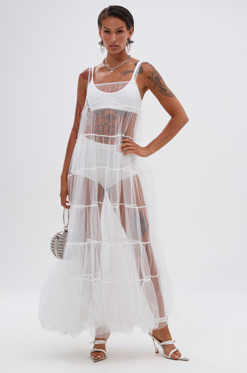 A person wears the PERFECT FOR LAYERING TULLE MAXI DRESS IN WHITE over undergarments, styled with white heeled sandals, a silver handbag, bold jewelry, and short hair—showcasing tattoos through the sheer design. 1