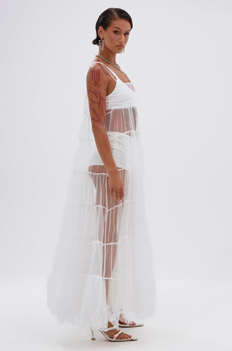 A person with slicked-back hair poses sideways in the PERFECT FOR LAYERING TULLE MAXI DRESS IN WHITE, styled over white undergarments and heeled sandals, with visible arm tattoos and silver jewelry against a plain white background. 4