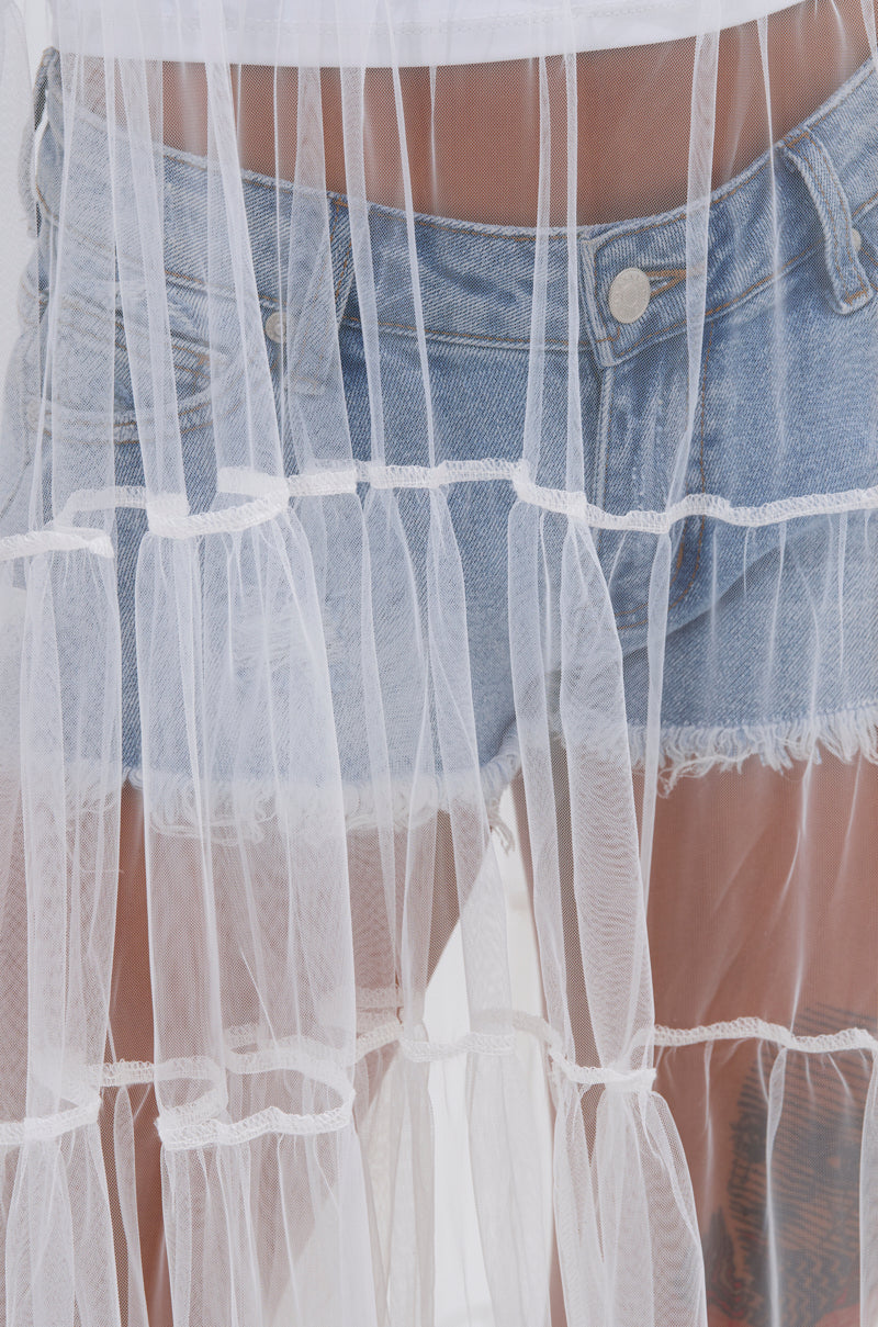A person in frayed denim shorts and a white top stands behind the PERFECT FOR LAYERING TULLE MAXI DRESS IN WHITE, with the sheer, pleated fabric revealing their lower torso and upper thighs. 6