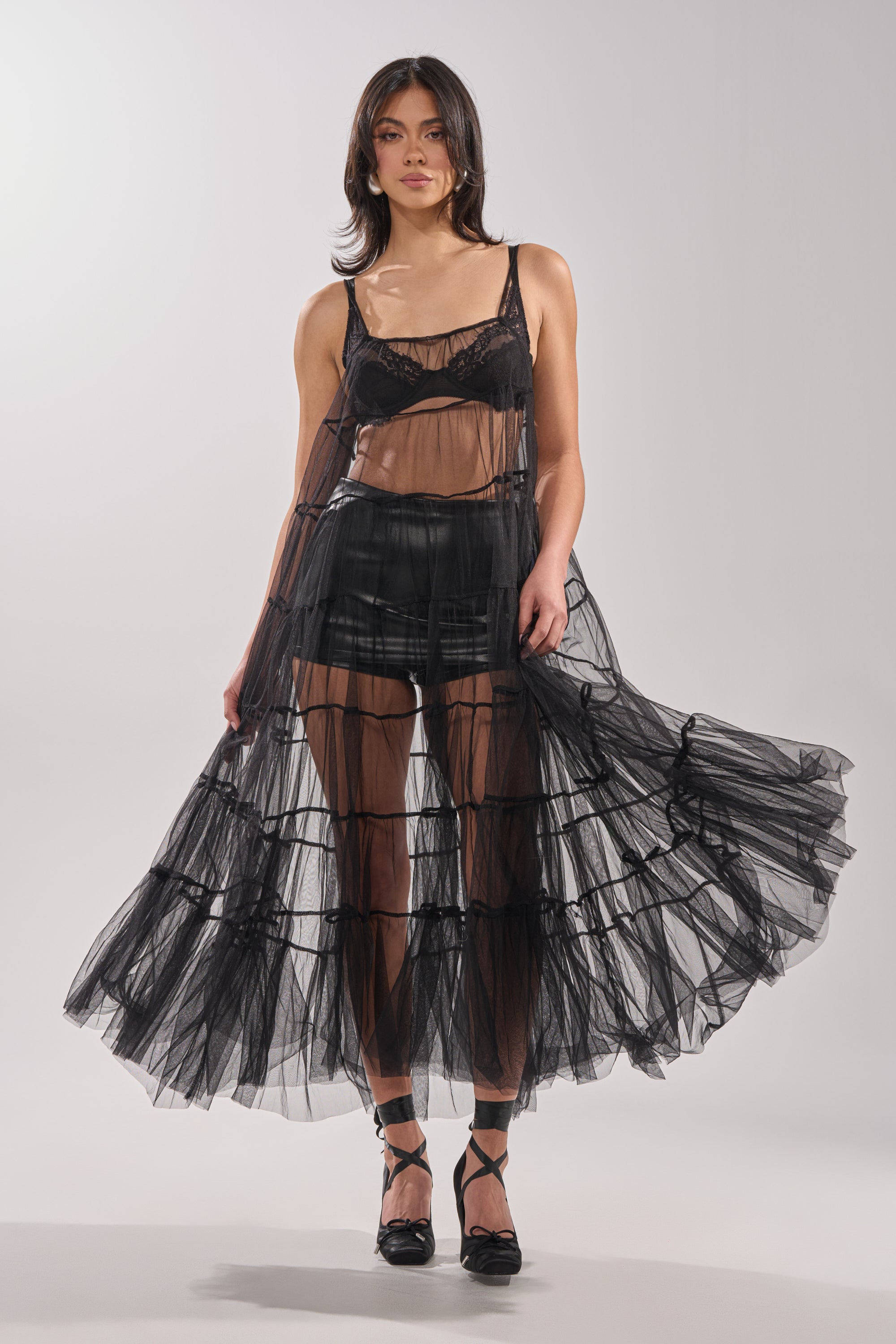 A woman models the PERFECT FOR LAYERING TULLE MAXI DRESS—a sheer, flowing black maxi dress styled over black lingerie and high-waisted shorts with strappy heels, against a simple light background. 0