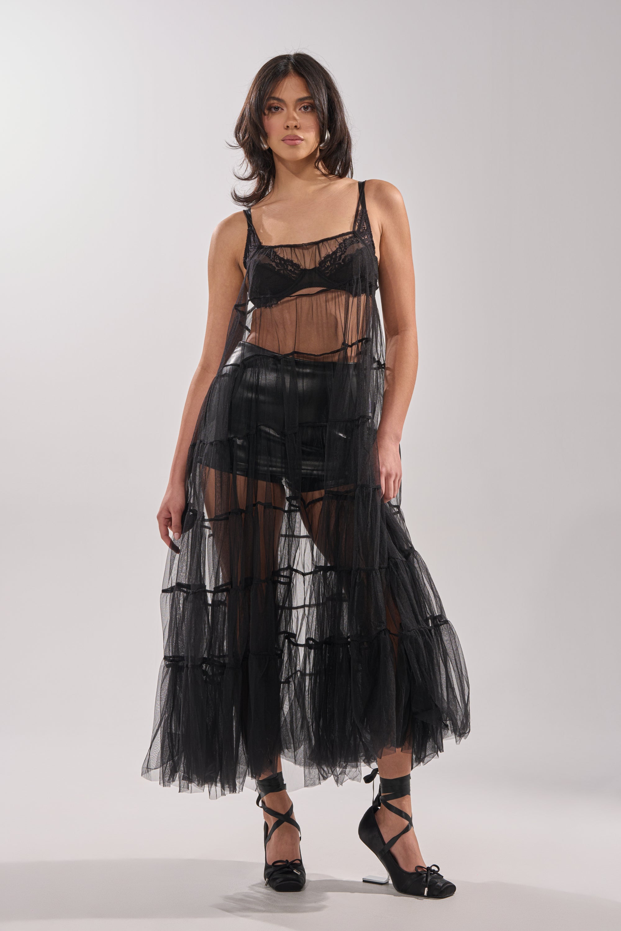 A woman with shoulder-length hair confidently poses against a plain backdrop in the PERFECT FOR LAYERING TULLE MAXI DRESS, styled over black shorts and a matching bralette with black strappy heels. 1