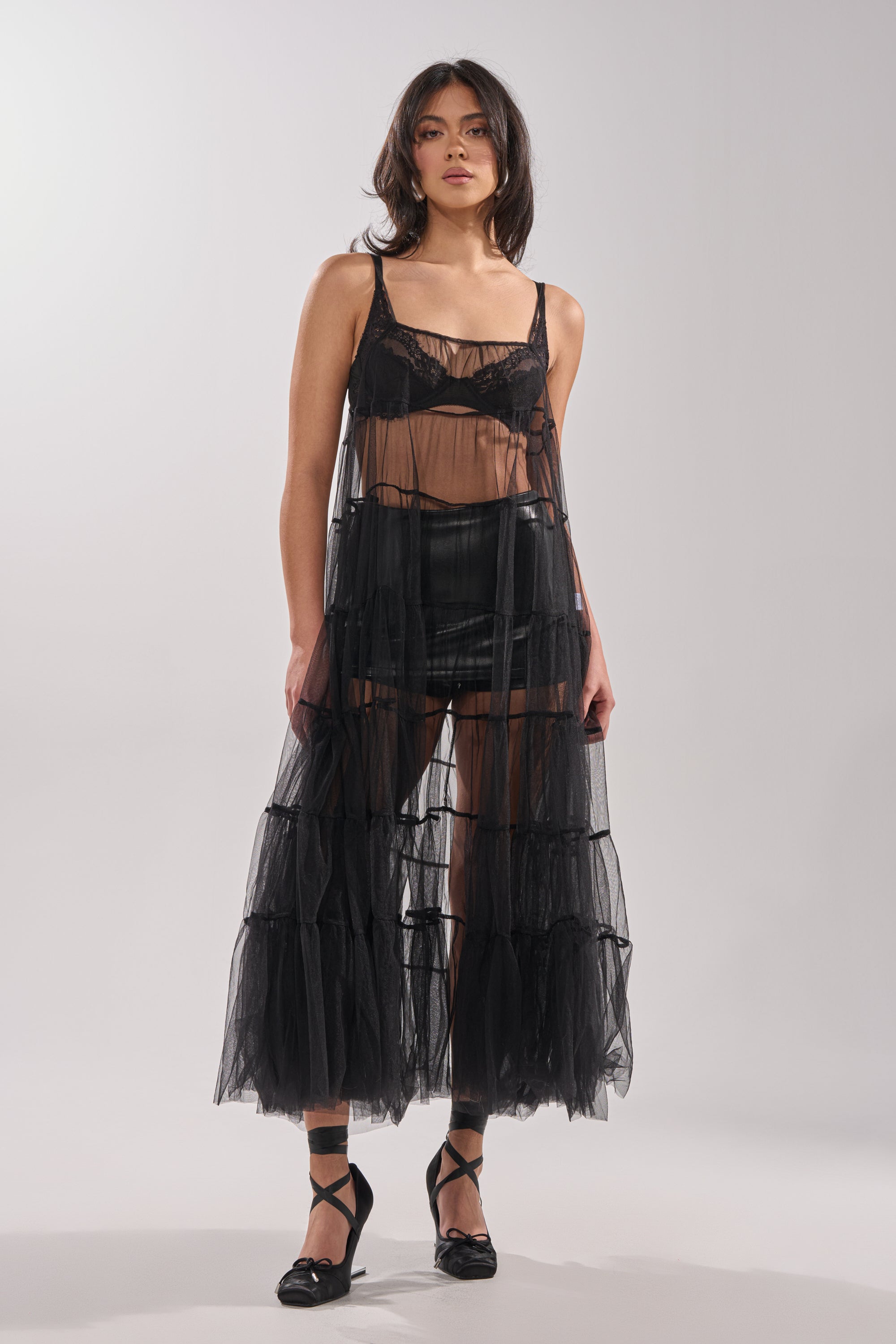 A woman with dark, shoulder-length hair poses against a plain background wearing the PERFECT FOR LAYERING TULLE MAXI DRESS over black lingerie and shorts, paired with black ribbon-tied ballet flats. 2