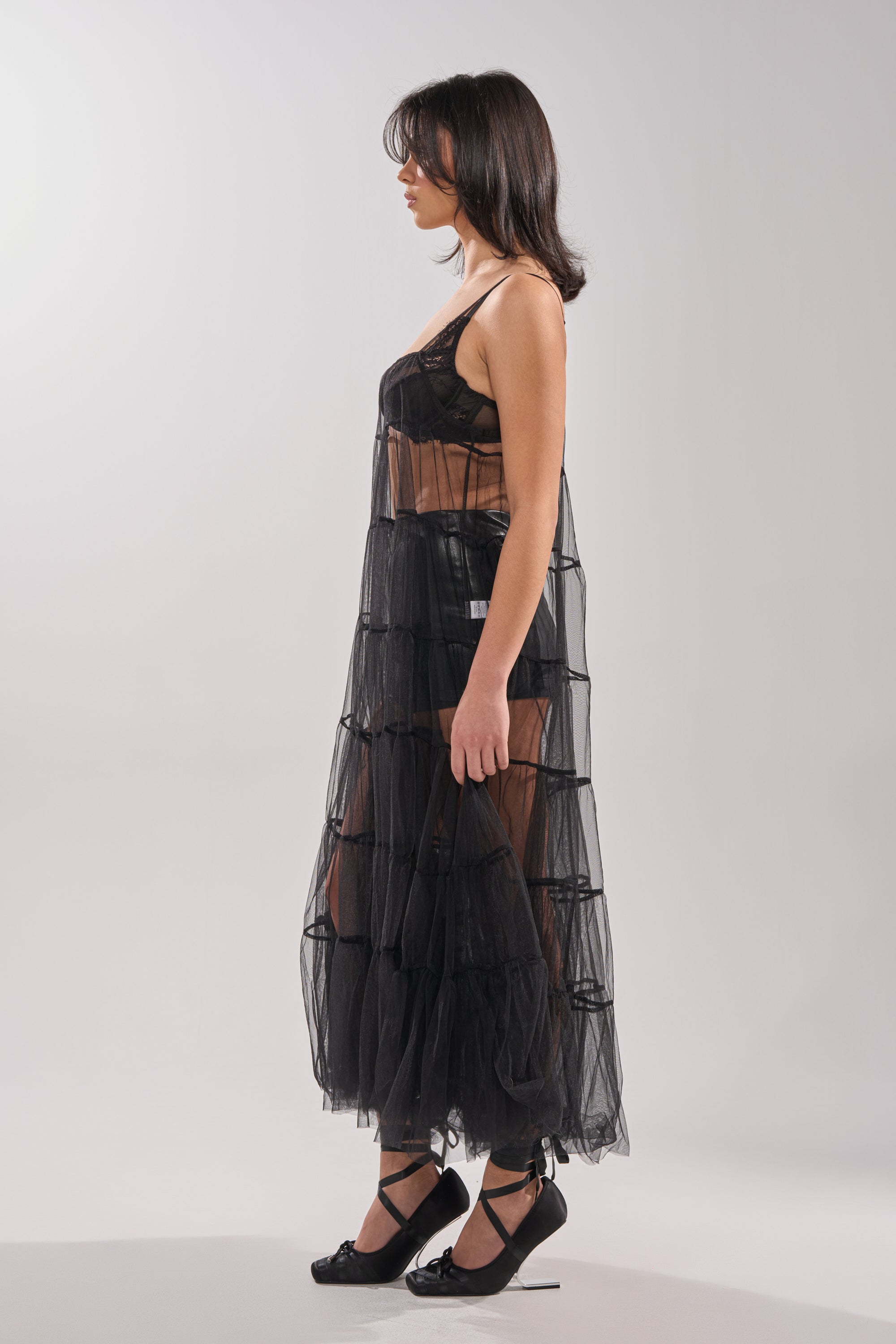 A woman stands in profile wearing the PERFECT FOR LAYERING TULLE MAXI DRESS—sheer, layered, and ruffled—over a black bralette and shorts with black heels, set against a plain light gray background. 3