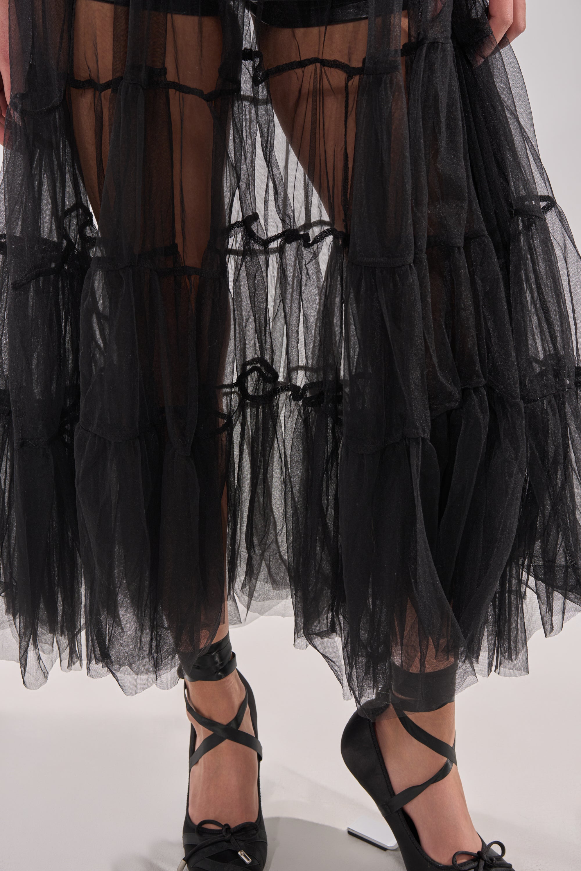 A close-up of a person wearing the PERFECT FOR LAYERING TULLE MAXI DRESS in sheer black, styled with black ankle-strap high heels featuring bows, standing on a light-colored floor. 6