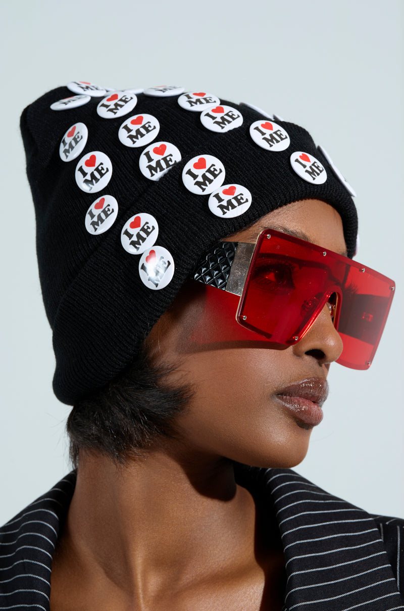 Wearing oversized red sunglasses and a black I REALLY DO BEANIE adorned with "I ❤️ ME" badges, a person completes their look with a black and white striped jacket. The side profile image showcases the beanie's stretchy knit, adding coziness to this statement ensemble.