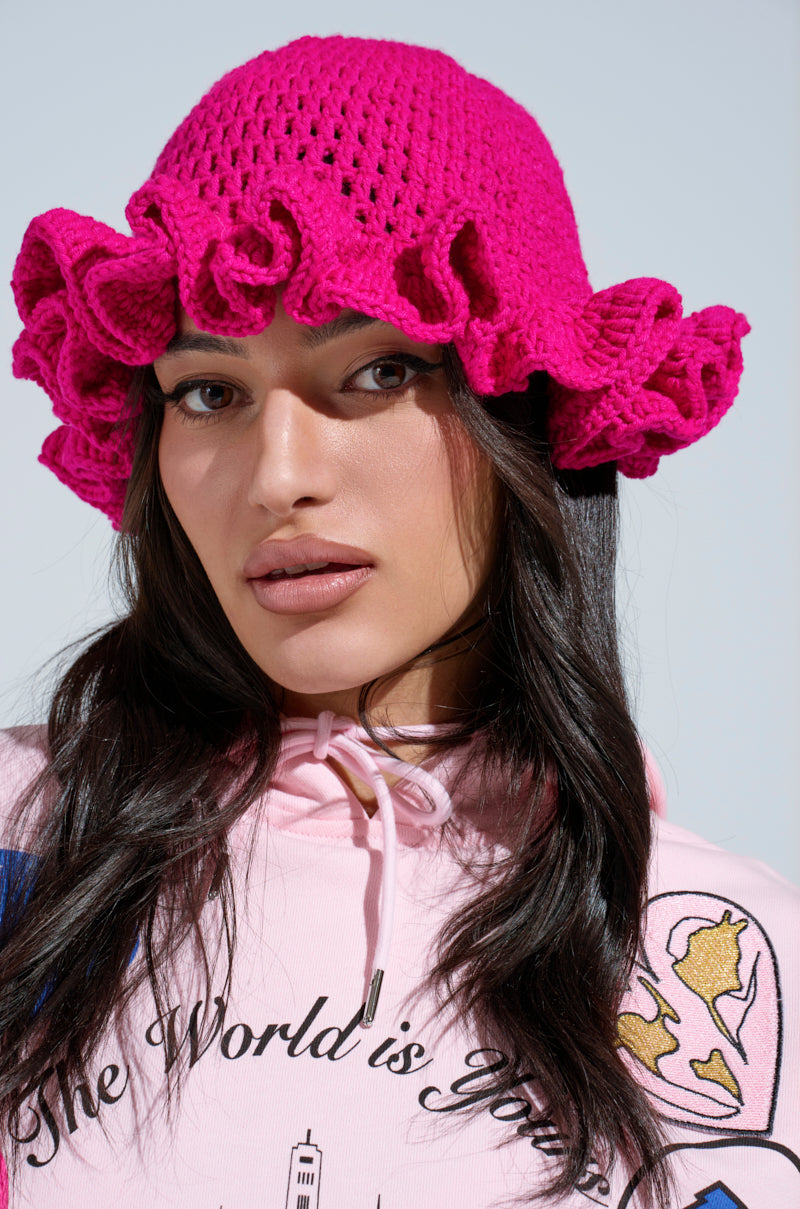 A woman with long dark hair wears the CROCHET NIGHTS HAT, featuring a bright pink hue and ruffled brim, paired with a light pink "The World is Yours" hoodie with globe and city graphics. She looks directly at the camera.