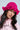 A woman with long dark hair wears the CROCHET NIGHTS HAT, a bright pink crochet hat with a ruffled brim, and a pale pink hoodie with black text, looking directly at the camera against a light background.